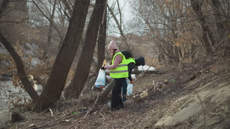 two volunteers wearing reflective safety vests and gloves picking up trash and debris from riverbank, placing waste into blue bags during outdoor cleanup to protect environment, reduce pollution