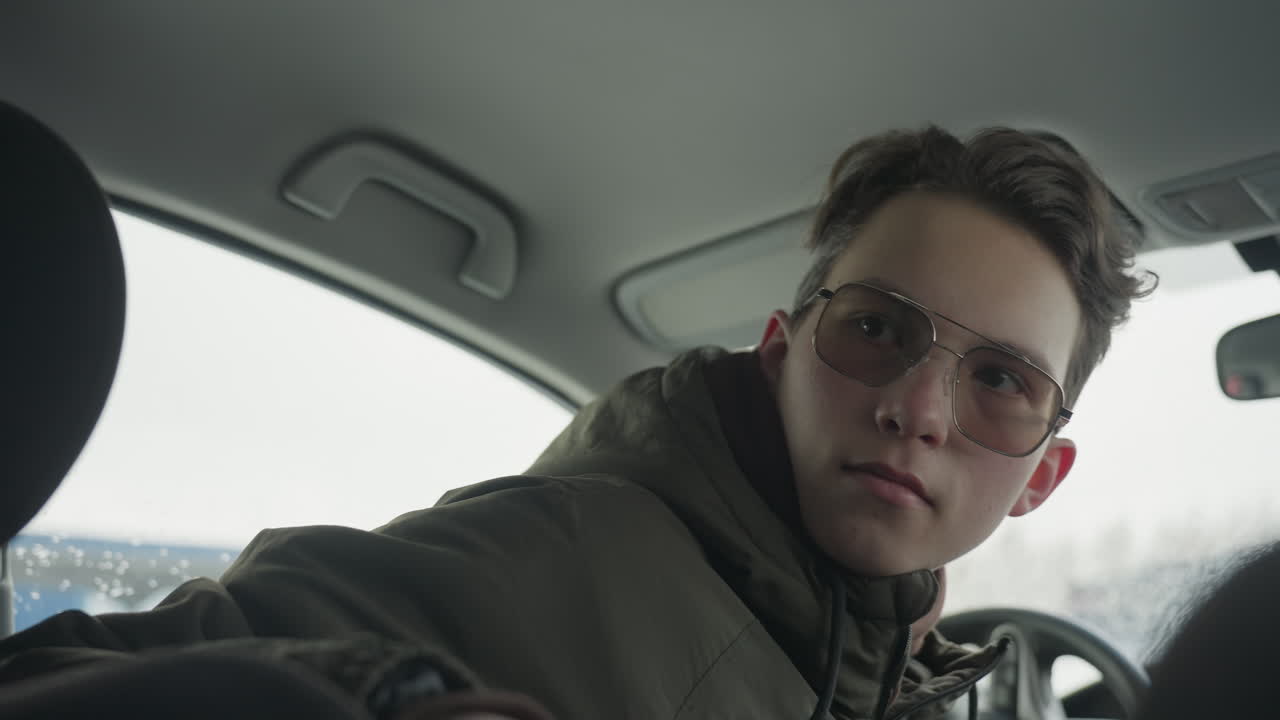 teenage boy in sunglasses and winter jacket sits behind steering wheel in parked vehicle, turns head back with subtle smile and looks directly at camera with light through rainy window background