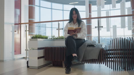 banker seated on stylish wooden bench in spacious modern mall reads book while flipping page, wearing formal white shirt and black trousers, surrounded by natural lighting