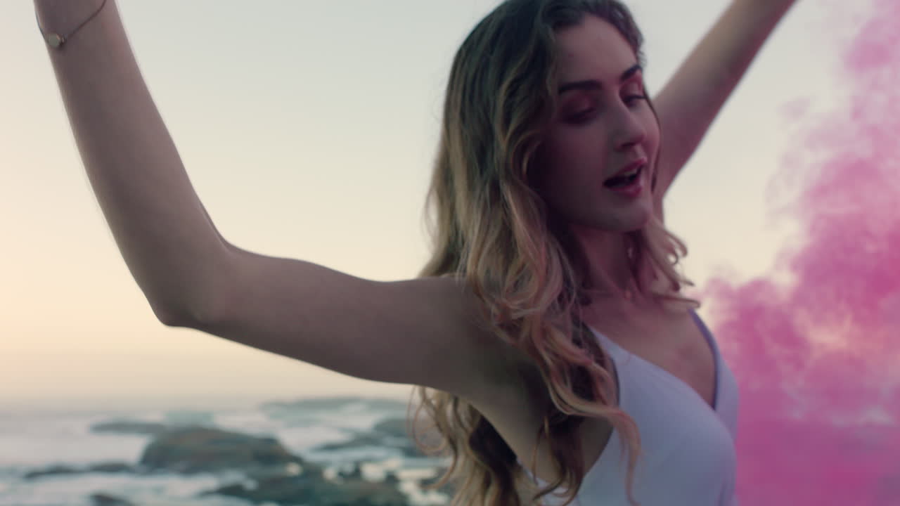 아름다운 여성이 분홍색 연기 폭탄을 흔들며 해가 뜨면 해변에서 춤을 추고 창조적 자유를 축하합니다.