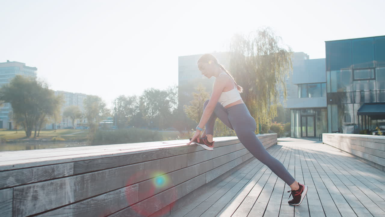 Athletic young woman runner stretching legs before jogging on bridge in city park on sunny day