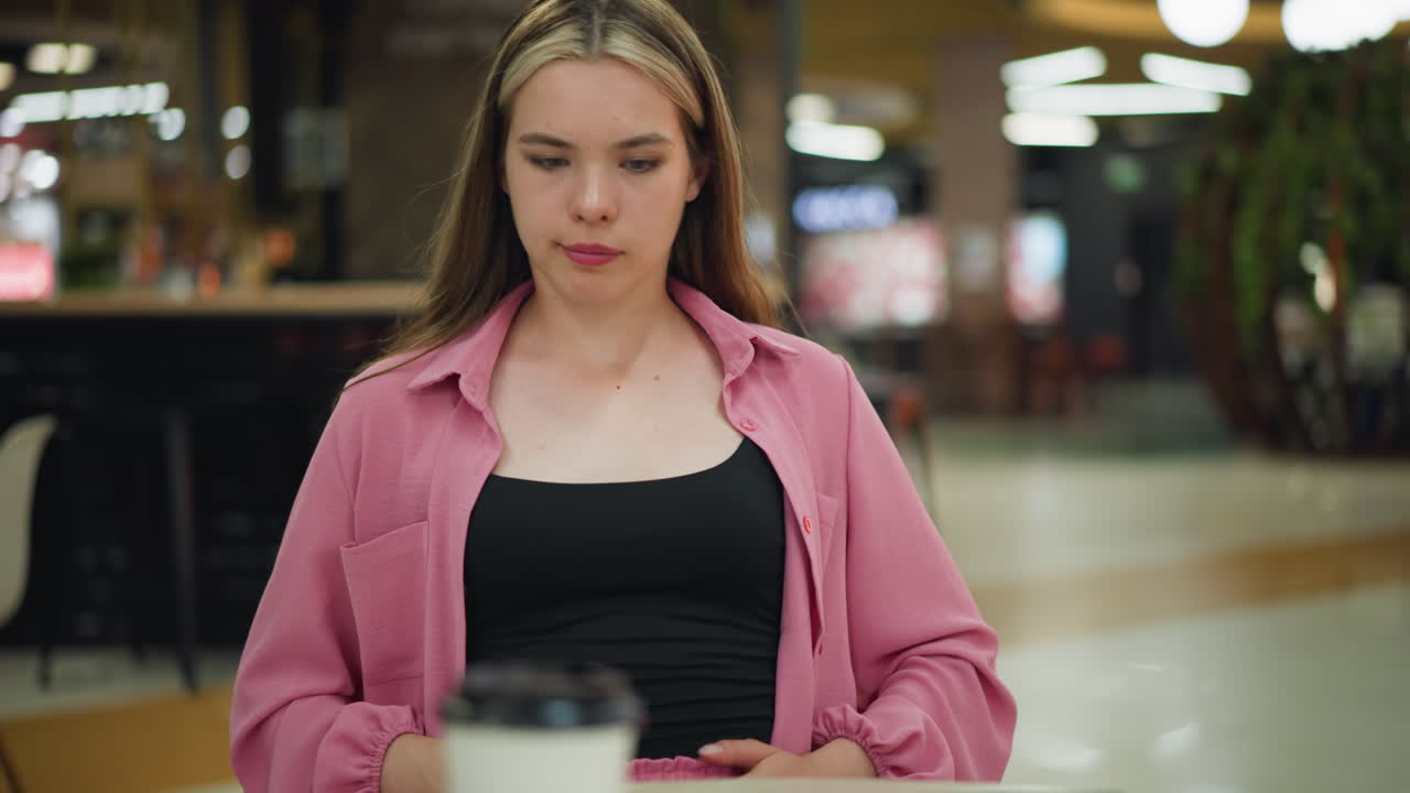 분홍색 드레스를 입은 여자가 식사 후 양손으로 배를 부드럽게 문지르고, 눈에 띄게 만족감을 느끼고, 커피 컵, 킨, 잔류 된 감자 김이있는 트레이가 그녀의 앞 테이블에 놓여 있습니다.