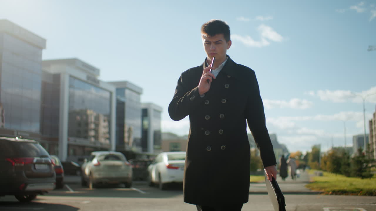 검은 코트를 입은 사업가는 전화를 계속하는 동안  에 전화를 들고, 주차된 자동차와 현대 건물 근처의 도시 환경에서 밝은 빛 아래 밖에서 자신감 있게 고 있습니다.