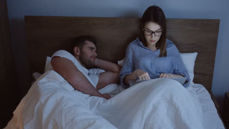 Handsome-young-man-in-pajama-and-glasses-working-late-at-night-on-the-laptop-computer-while-his-wife-sleeping-peacefully-near-him-in-the-bed