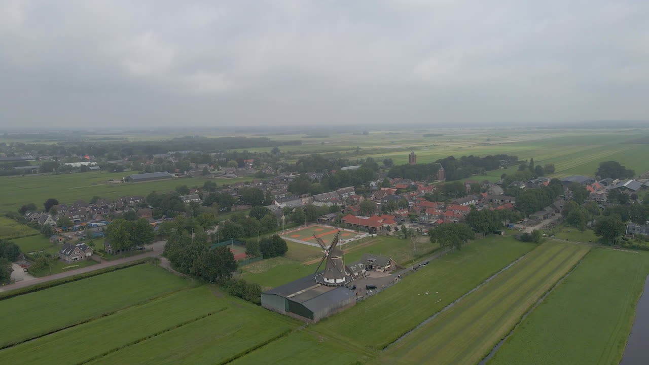 녹색 초원으로 둘러싸인 회전하는 오래된 풍차가 있는 작은 시골 마을을 향해 날아갑니다.