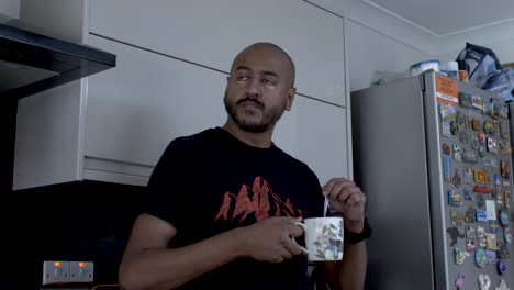 bald adult male indian stirring tea in mug, resting back on kitchen counter and looking right thinking