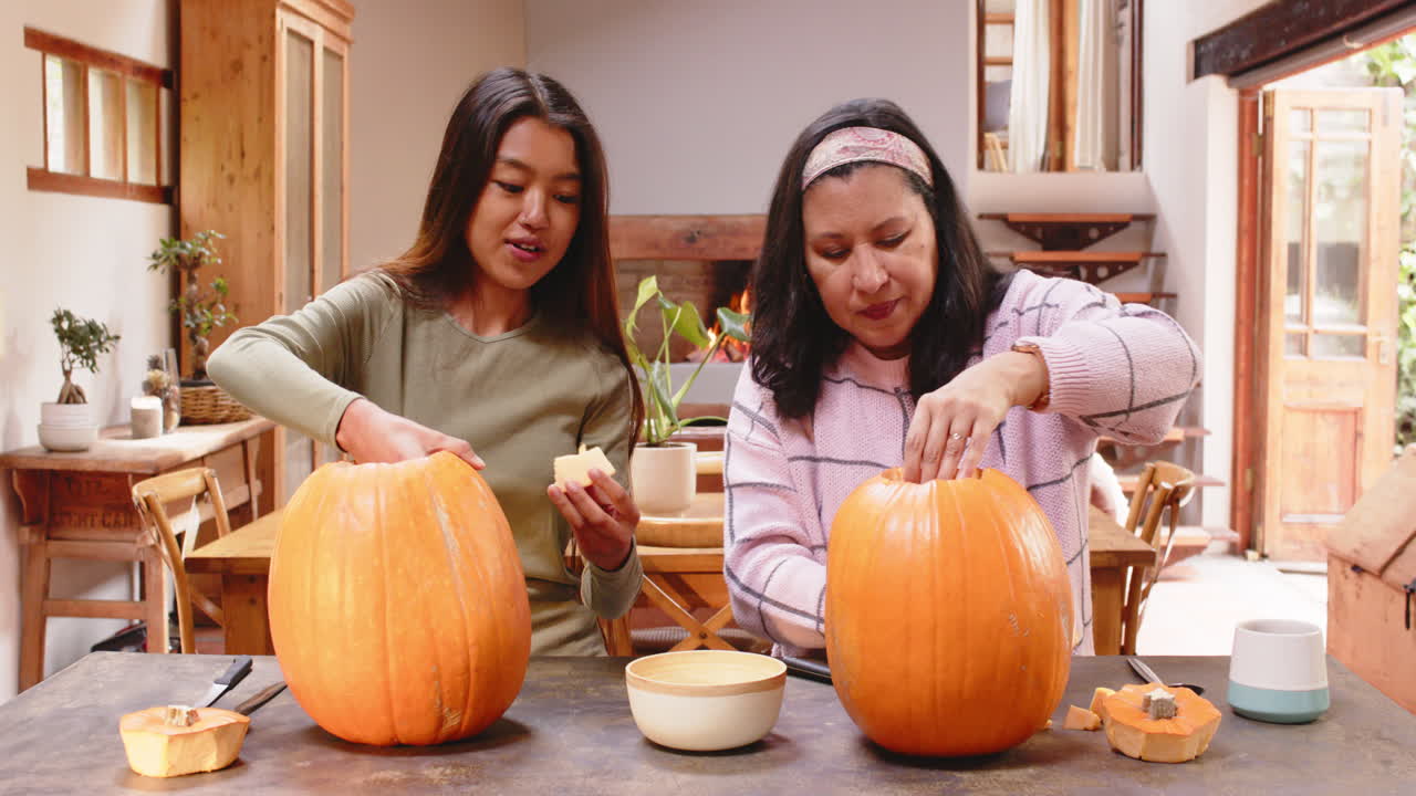 Carving pumpkins together, multiracial grandmother and young woman, at home, Halloween time