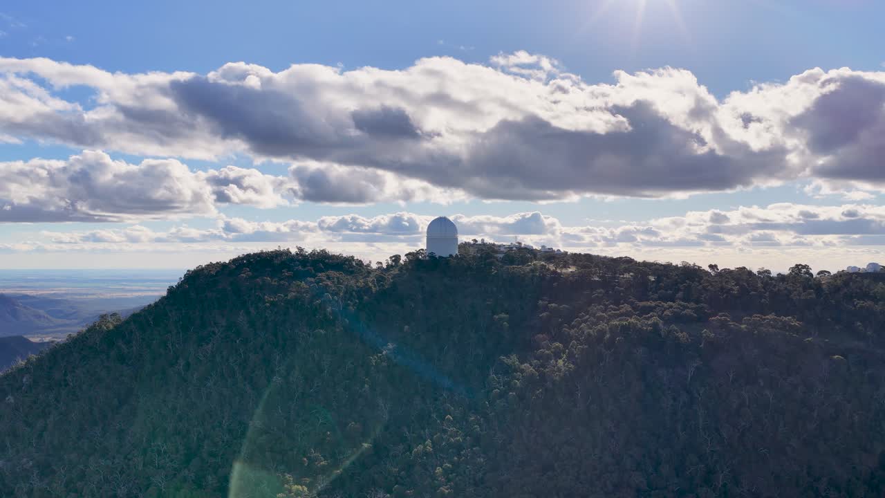 Aerial drone footage smoothly pans across a forested mountain ridge with a large observatory dome under dramatic morning sunlight and scattered clouds