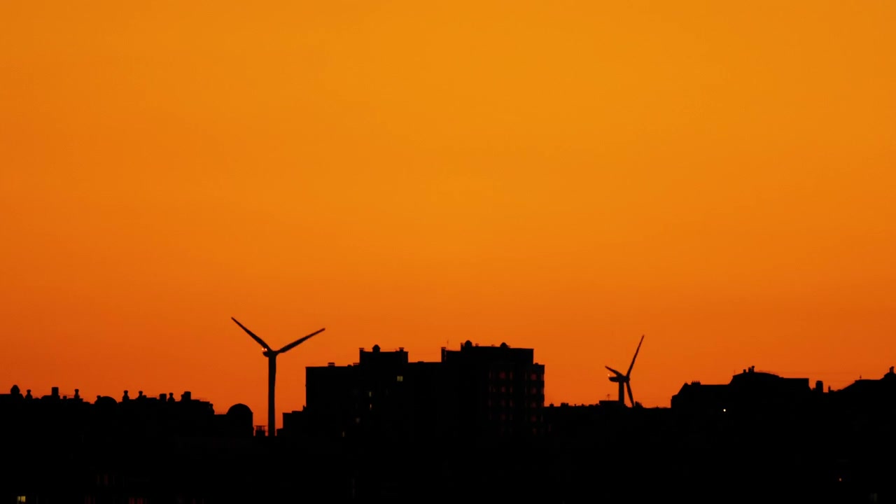 Lapso de tiempo de aerogeneradores silueteados girando sobre el horizonte de una ciudad durante un atardecer dorado