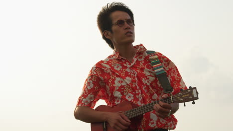 low angle shot of an asian man standing on top on a mountain, singing a song while playing ukulele on a cloudy day