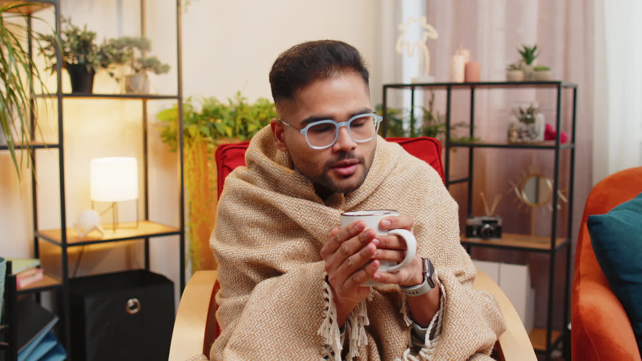 Sick indian man wrapped in plaid sit alone shivering trembling from cold on chair drinking hot tea