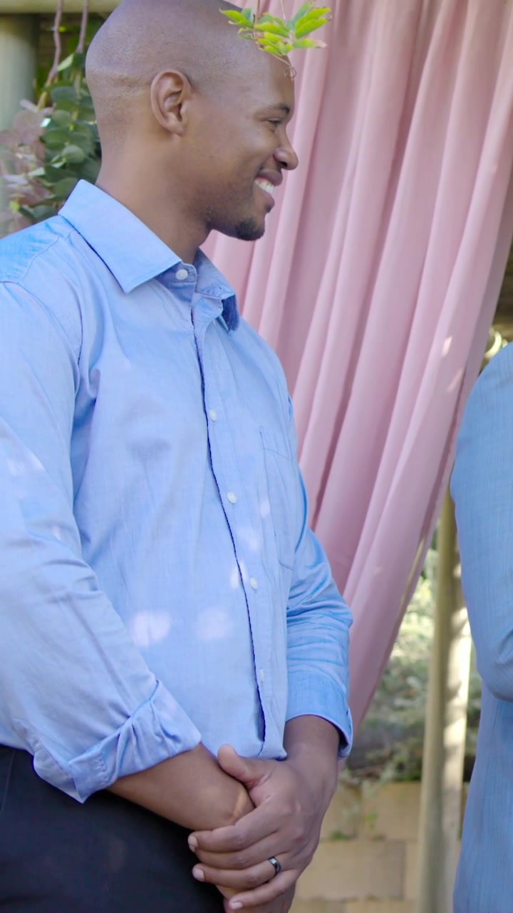Video verticale: Uomo in camicia blu che reagisce al segnale dell'altoparlante, girando la testa e sorridendo in giardino, con un drappo rosa.