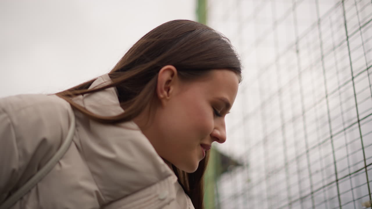Primo piano di una giovane donna sorridente attraverso una recinzione, delicata visuale di profilo con giacca leggera, sfondo a rete che suggerisce un campo sportivo, espressione allegra e postura inclinata, atmosfera da spettatrice spontanea, attenuato