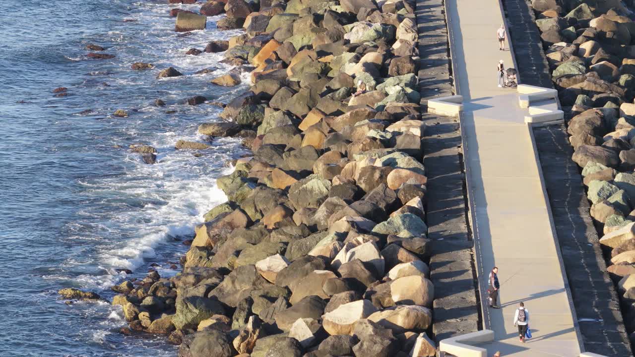 Drone captures people walking along a sunlit pier surrounded by ocean waves and rocky barriers