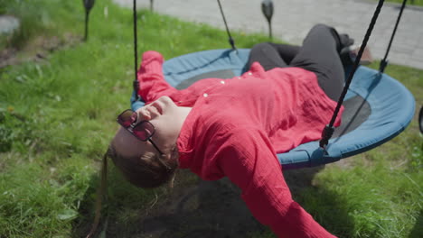 smiling woman in sunglasses and red shirt enjoys peaceful moment lying on circular outdoor swing on sunny day, surrounded by green grass, she relaxes freely with arms stretched