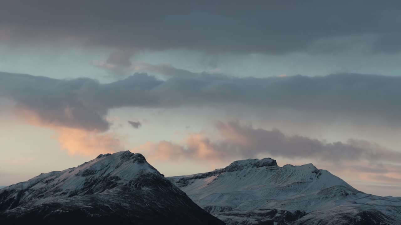 Pastel coloured clouds over snow covered mountain peaks at dusk, telephoto time lapse