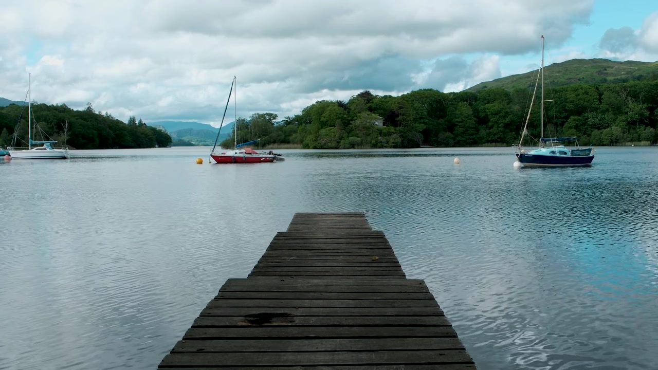 Inquadratura statica da un pontile di legno attraverso il lago Coniston nel Lake District, che rivela barche a vela ormeggiate, riflessi calmi, boschi circostanti e colline lontane.