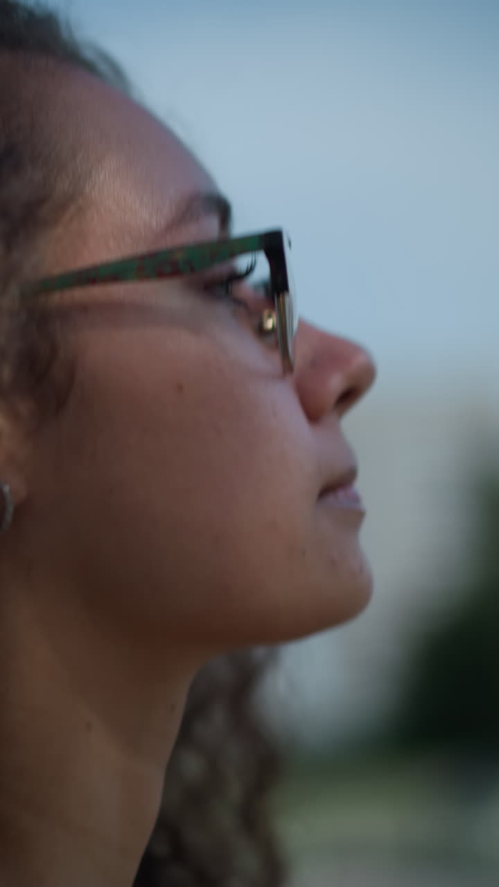 primer plano de una mujer con cabello rizado y gafas caminando al aire libre, ajustando el cabello hacia atrás mientras la luz del sol proyecta sombras suaves en la cara, con vista borrosa de personas y edificios en el fondo