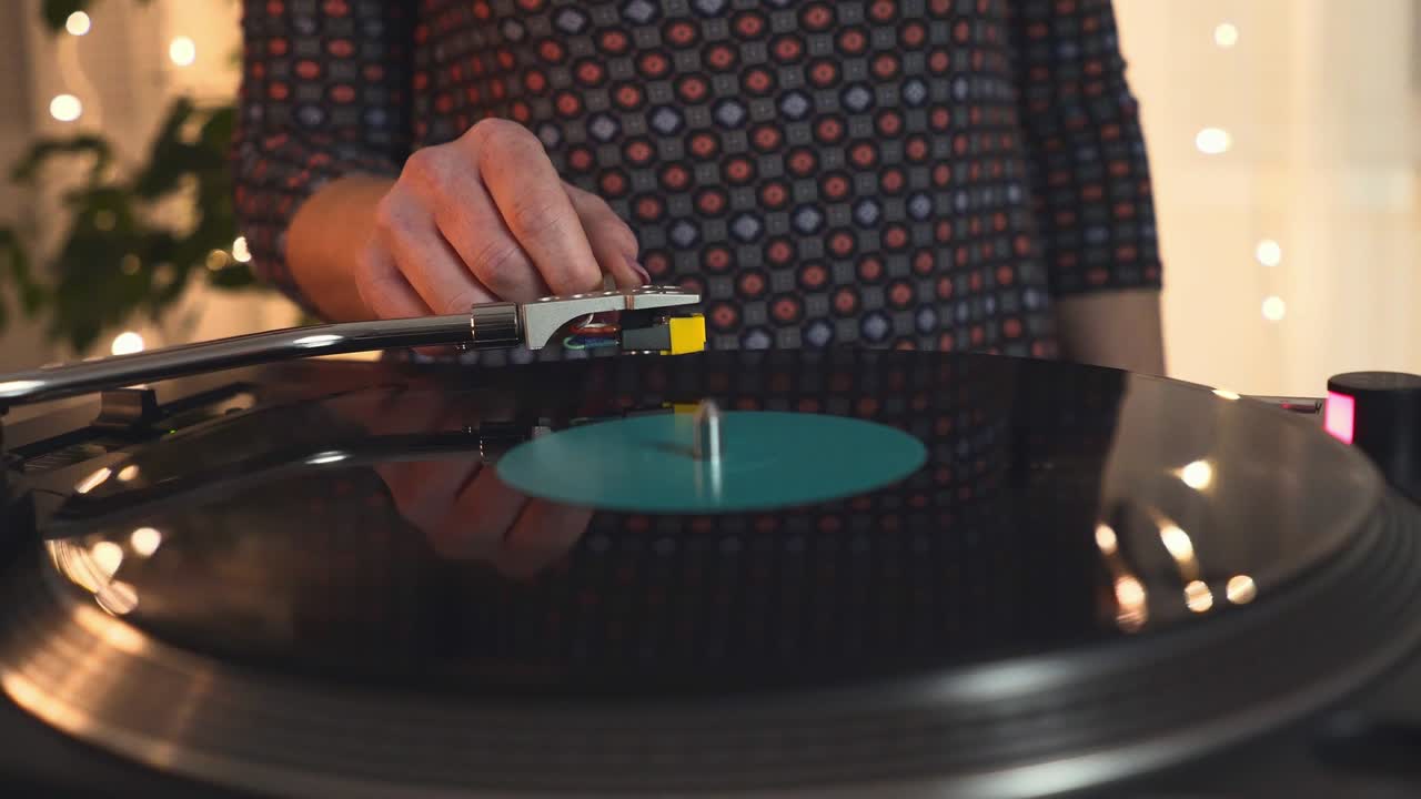 드레스를 입은 한 소녀가 dj 턴테이블 레코드 플레이어에 가까이 다가와 회전하는 검은 비닐 레코드에 바늘을 가진 스타일러스를 넣고 밝은 파티 조명을 배경으로 춤을 추고 으로 움직입니다.