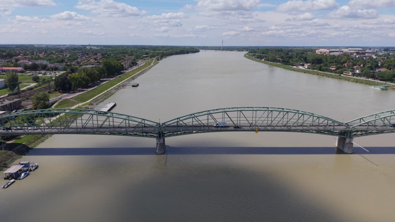 A green steel arch bridge spans a wide, calm river, with vehicles crossing in both directions. The scene includes riverside walkways, residential areas, and a modern bridge visible on the horizon