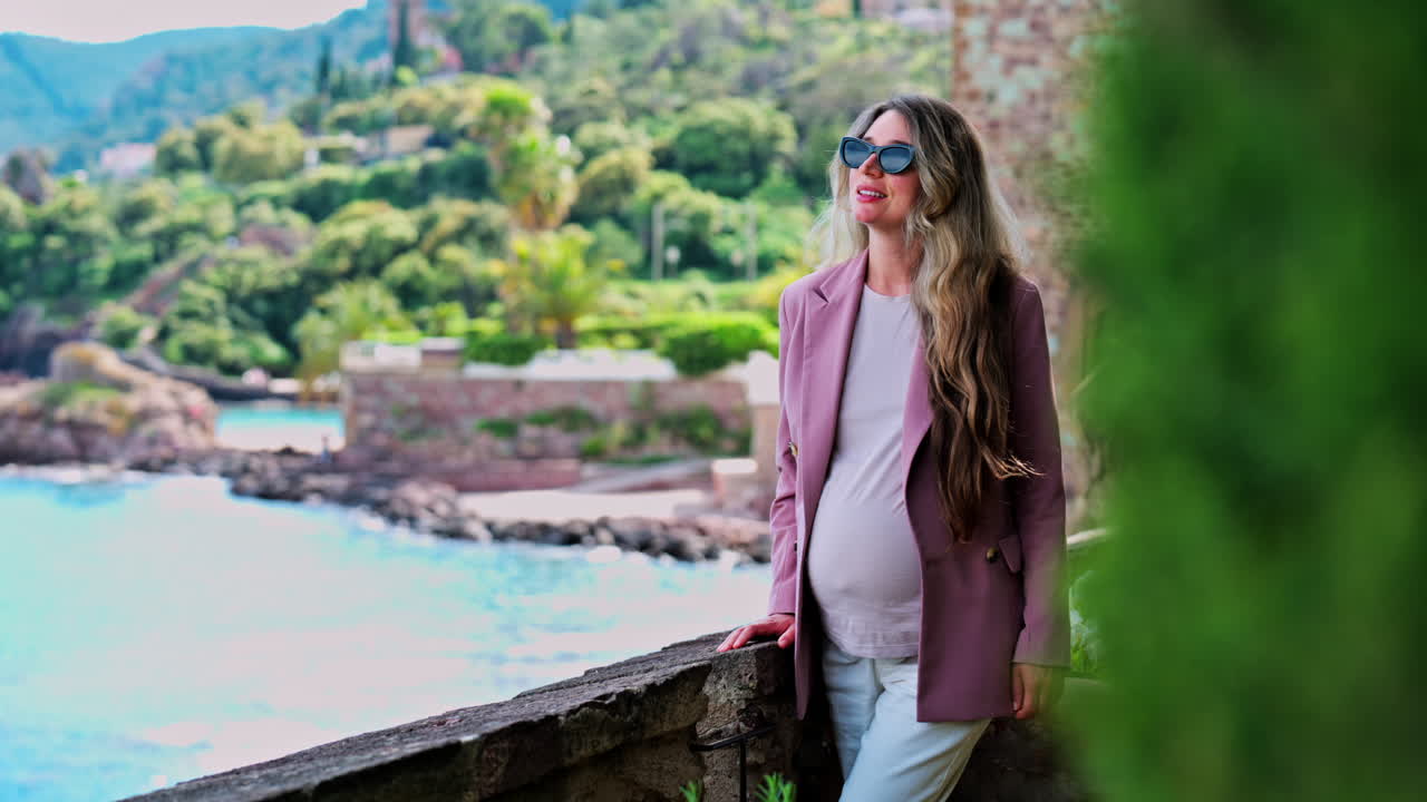 Pregnant woman with long hair, wearing sunglasses standing with the Chateau de la Napoule Castle in Mandelieu-La Napoule, France in the background
