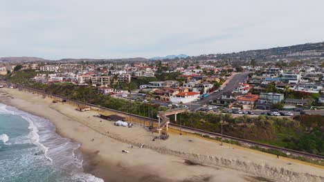 Vista-Aérea-De-La-Ciudad-Costera-De-California-Con-Surfistas-Cruzando-El-Puente-Sobre-Las-Vías-Del-Tren.