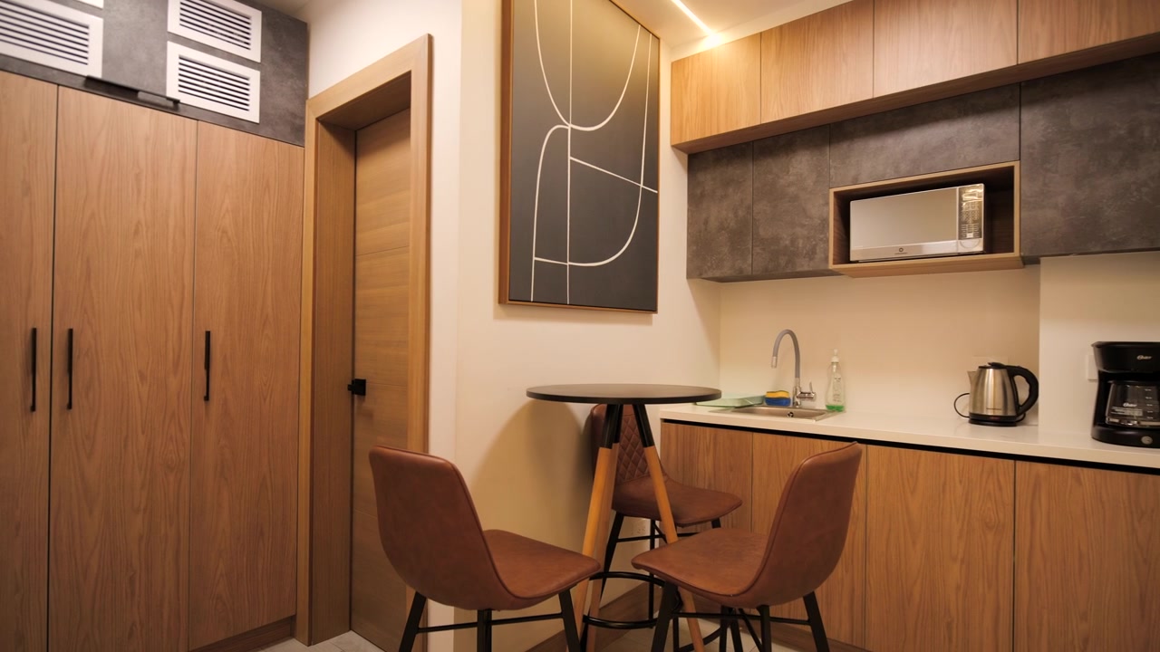 Inquadratura completa di uno spazio interno all'interno di un ufficio: arredamento in legno combinato, zona preparazione con lavello, microonde, macchina da caffè e bollitore, tavolo alto con sedie e opere d'arte astratte decorative.