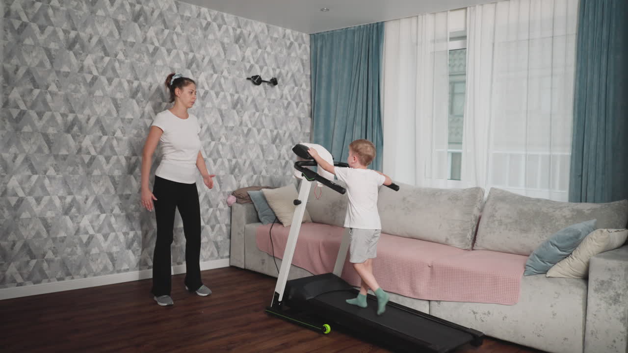 Young boy stands on treadmill confidently pressing control panel while female instructor in casual sportswear walks over curiously to observe his actions in bright cozy living room setting