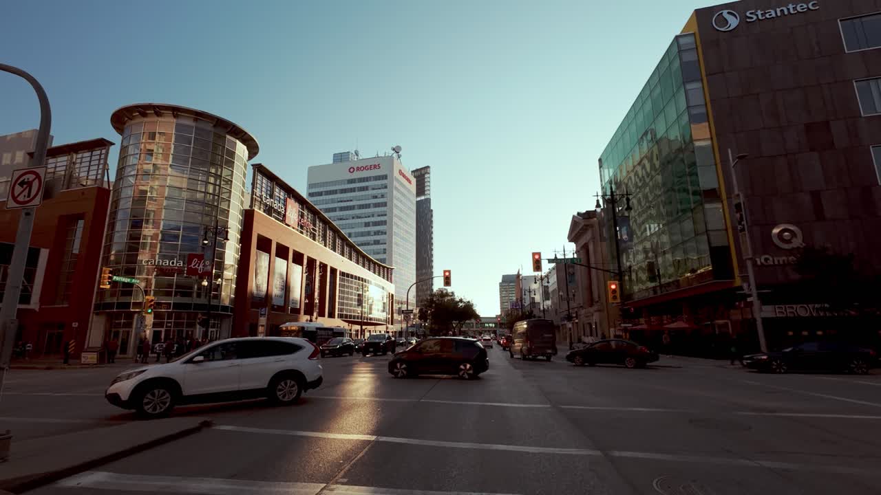 POV shot from car stopped at stoplight near Canada Life Centre; fans in jerseys cross street at golden hour.