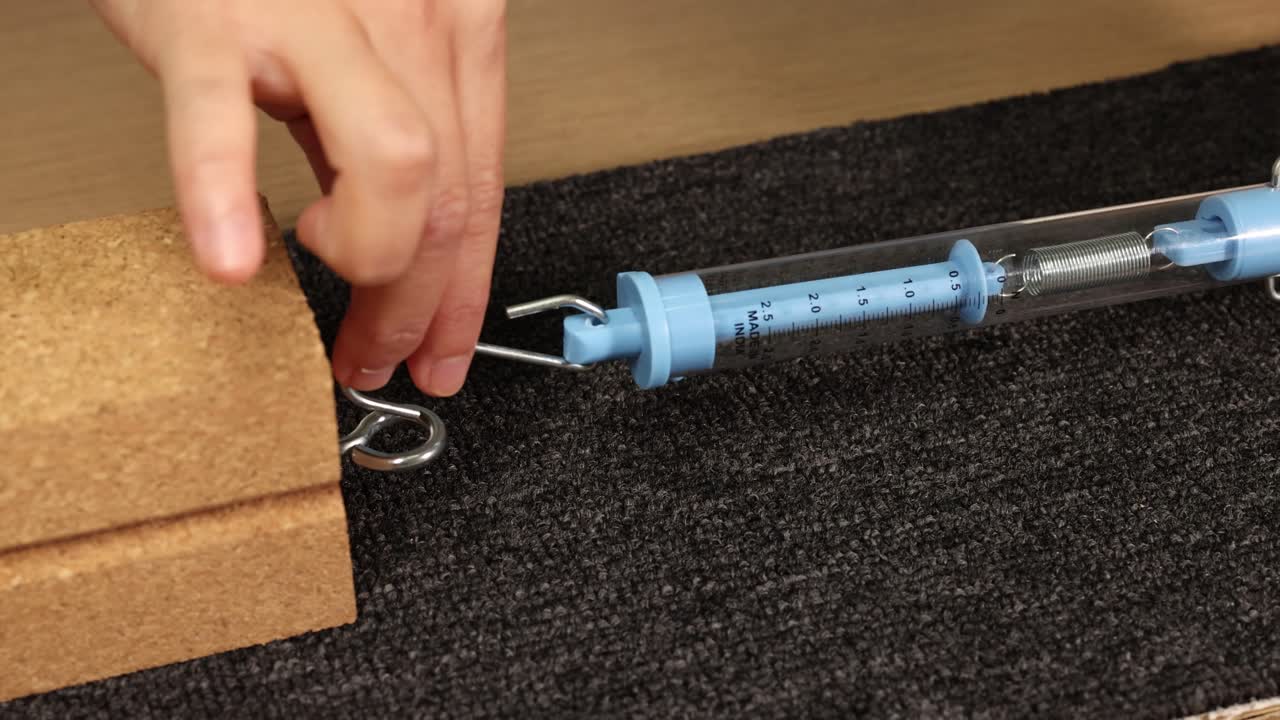 A hand uses a spring scale to measure friction between a wooden block and surface. Close-up, macro view with natural lighting