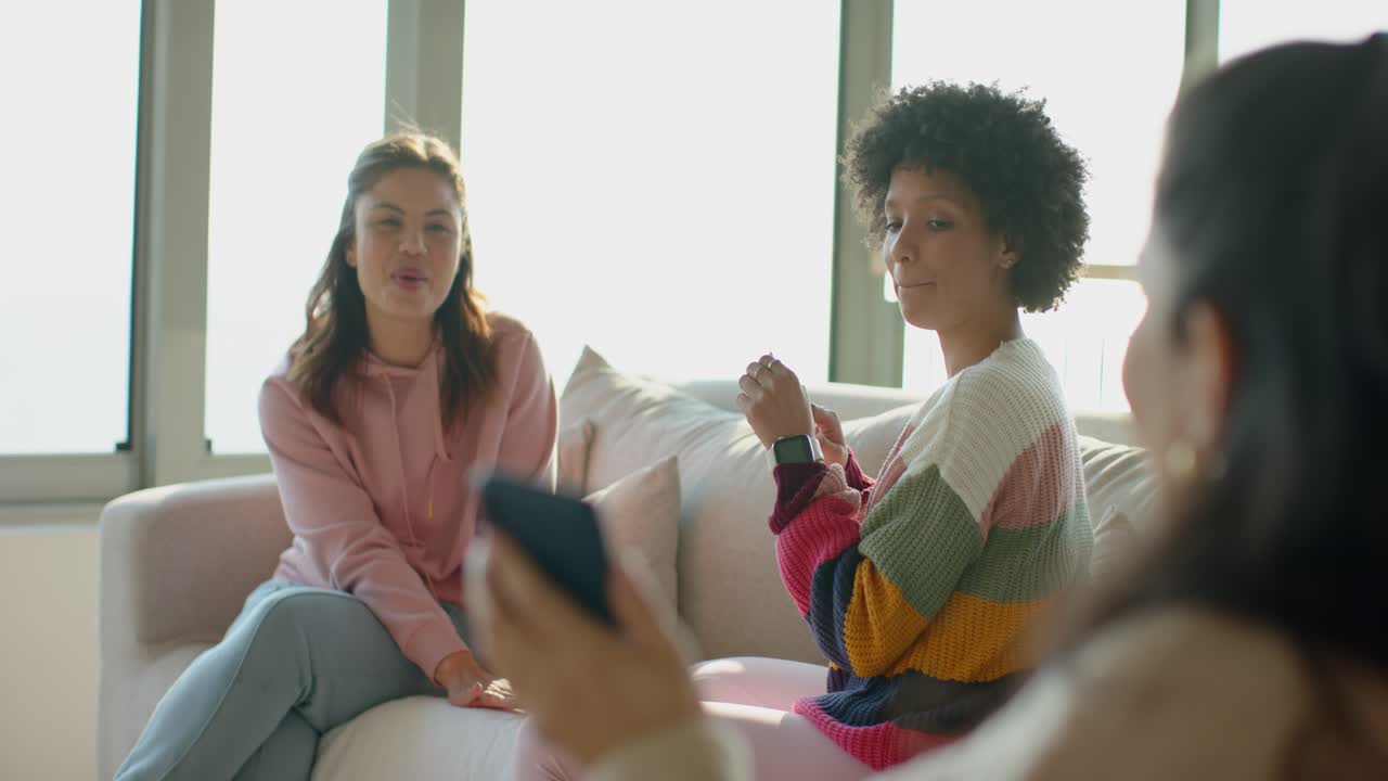 Diverse female friends laughing while third friend entering from right sharing phone screen on sofa