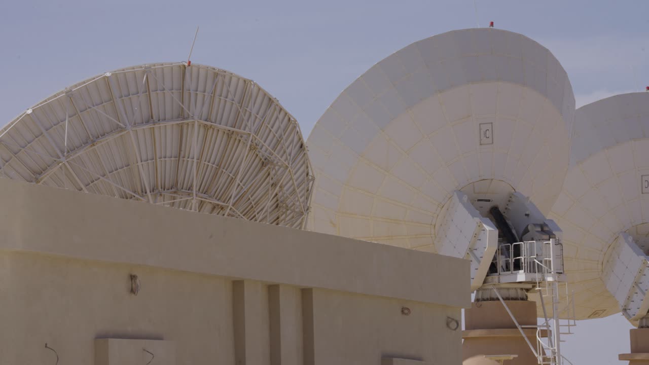 A static shot showcasing multiple generations of satellite technology. A large, complex mesh dish stands beside modern, solid parabolic antennas at a ground station