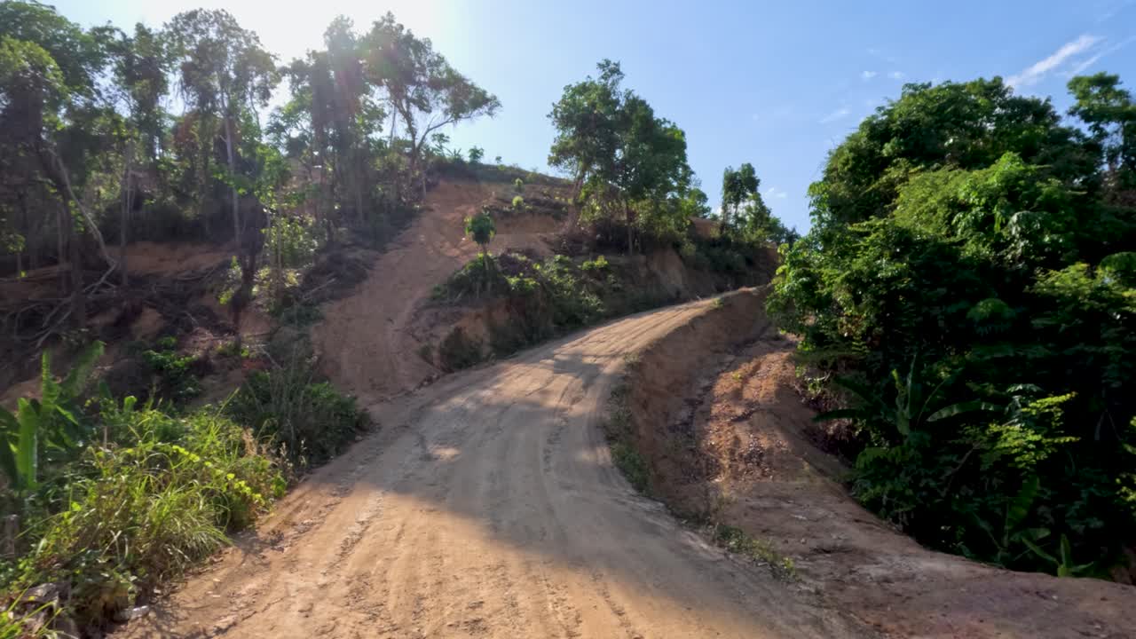 Vehicle travels a sunlit, rural dirt road through lush, hilly landscape with steady forward motion