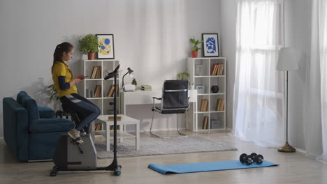 Una-Mujer-Está-Haciendo-Deporte-En-La-Sala-De-Estar-De-Su-Moderno-Apartamento-Sentada-En-Una-Bicicleta-Estática-Y-Usando-Un-Teléfono-Inteligente-Con-Un-Estilo-De-Vida-Saludable-Y-Una-Dama-Deportiva.