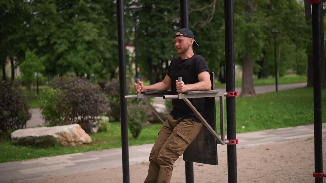 Outdoor L Sit Style Leg Raise On Metal Seat With White Male Holding Bars, Tight Core Engagement, Cargo Pants And Baseball Cap, Calm Park Surroundings And Concentrated Breathing For Abdominal Strength