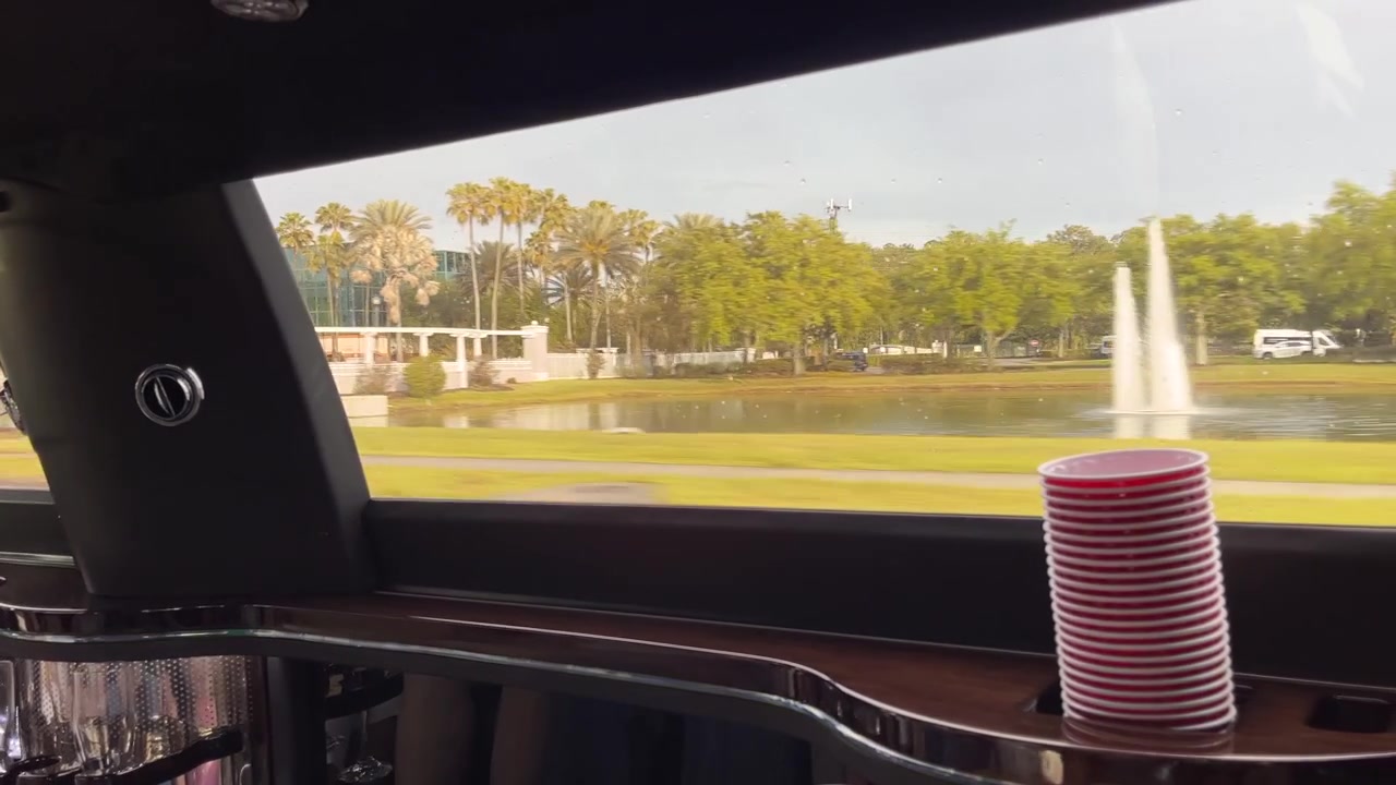 Janela de limusine emoldurando uma fonte de lago e palmeiras ao passar por um resort na Flórida. Interior de limusine de festa luxuosa com copos vermelhos com vista para a água e a estrada.