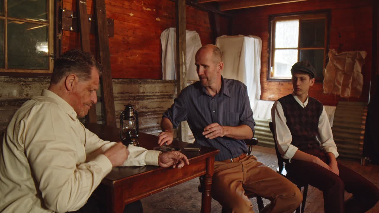 A man sits at a table in wearing 1930s style clothing. He smokes a cigarette, another man at the table asks for a cigarette. The man gives and lights the cigarette