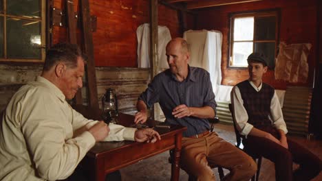 a man sits at a table in wearing 1930s style clothing. he smokes a cigarette, another man at the table asks for a cigarette. the man gives and lights the cigarette