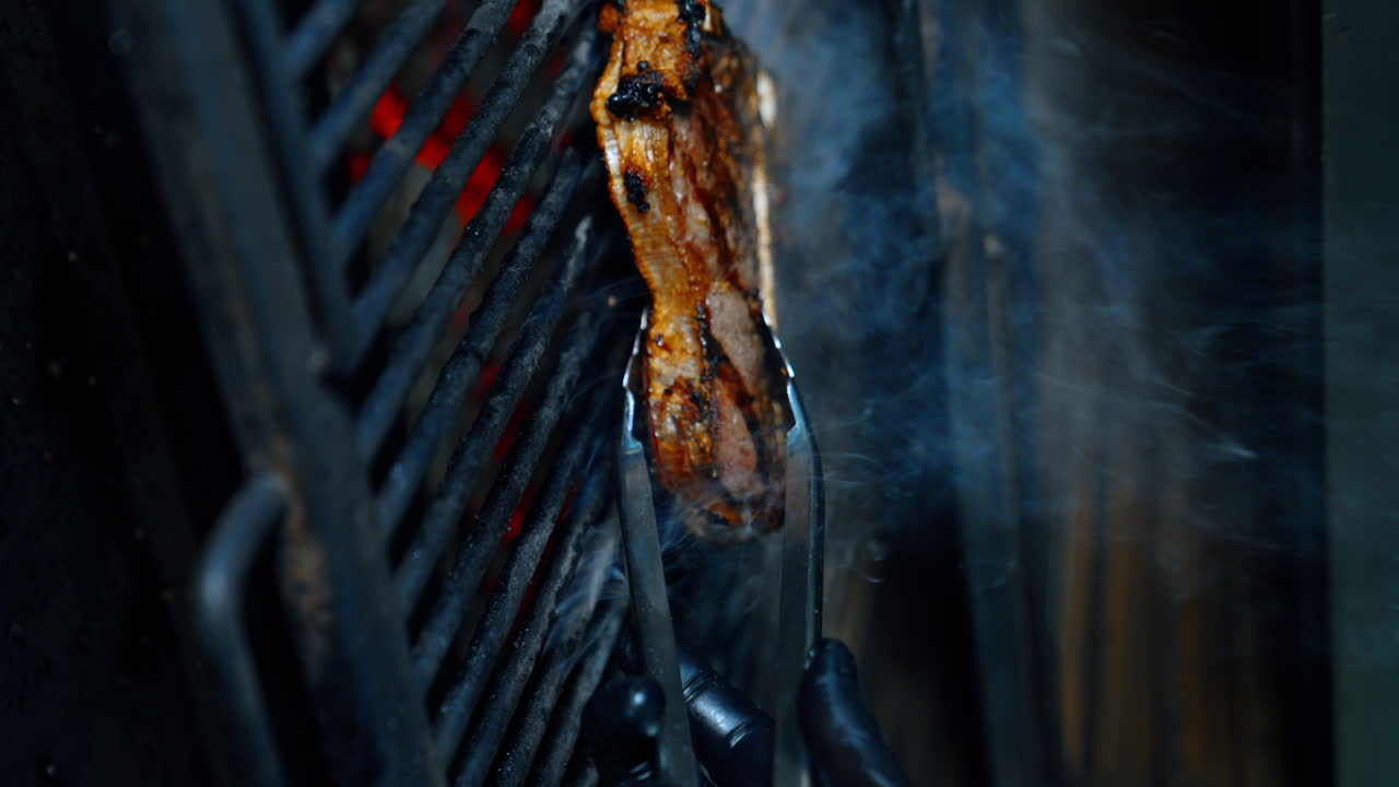 Meat on the bone is cooked on the grill. Gloved hand turns the piece of beef with tongs. Vertical screen. Close up.