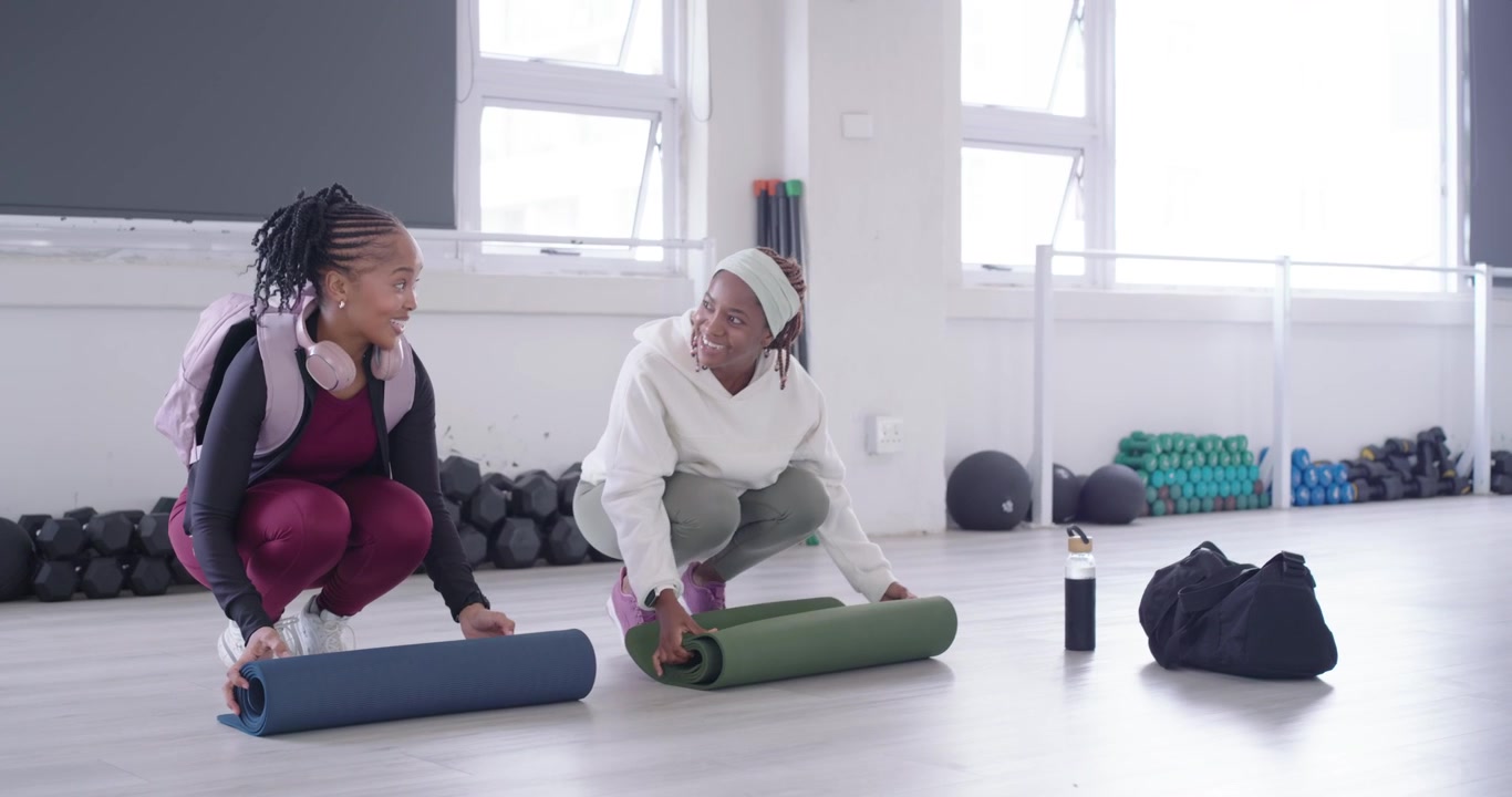 Two African women arriving at fitness studio carrying blue and green mats, unrolling for class