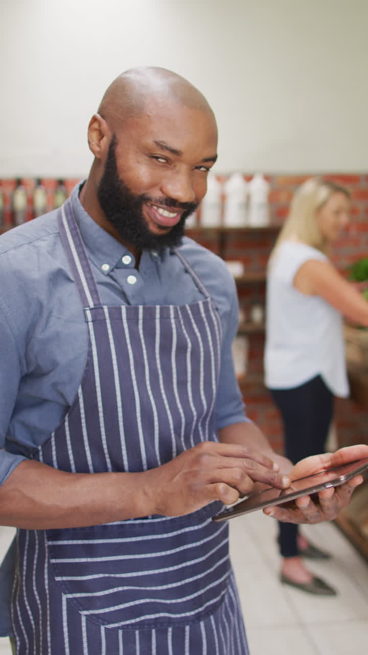 Vertical video of smiling african american in apron using tablet in organic store