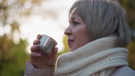 close up of lady outdoors in autumn light, wrapped in chunky knit scarf, lifting cup of steaming tea to lips and sipping slowly, expression calm and reflective against blurred foliage background