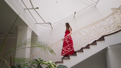 pretty woman in red gown goes down stairs. beautiful lady with brown loose hair wearing red slit dress comes to visit party in luxury residence