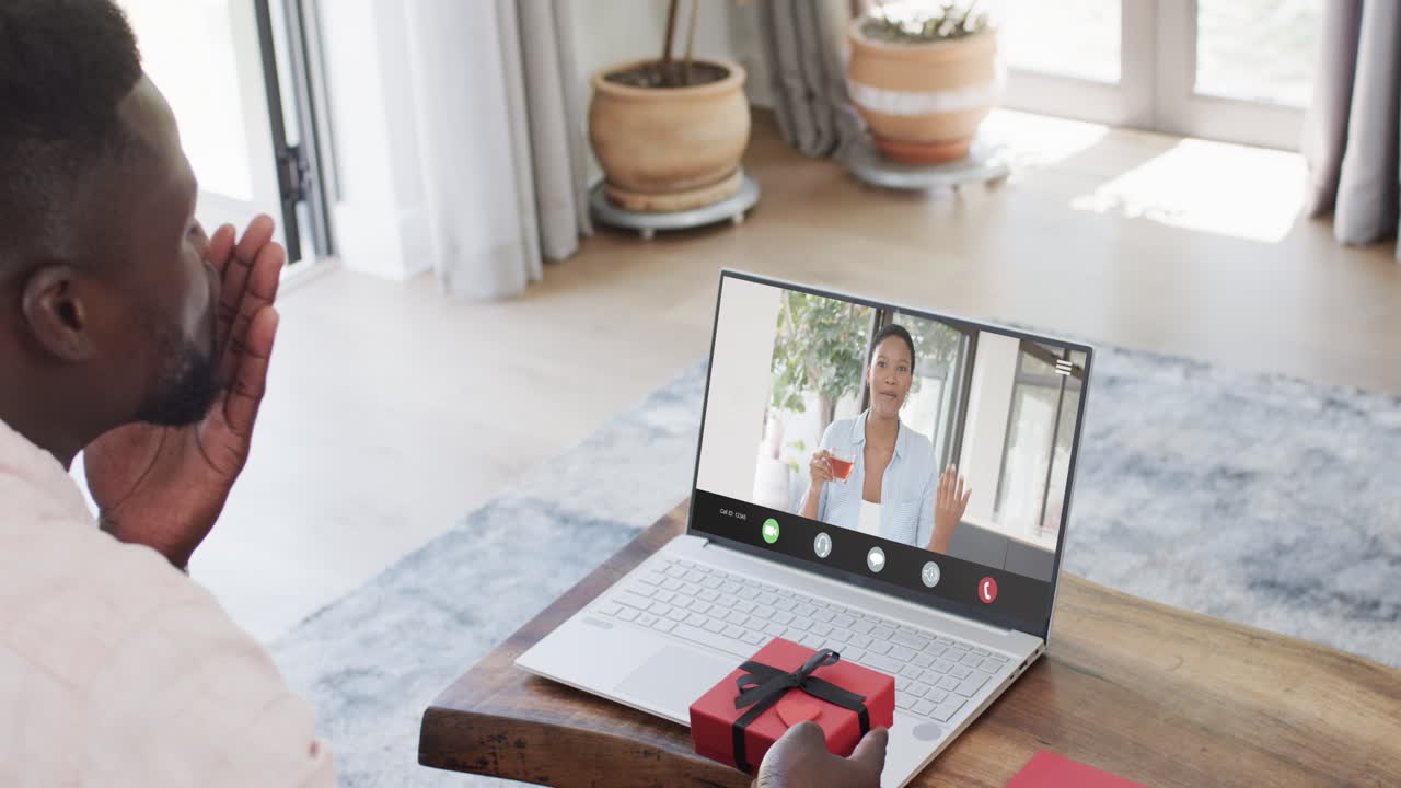 African american man holding red gift and using laptop with biracial woman on screen