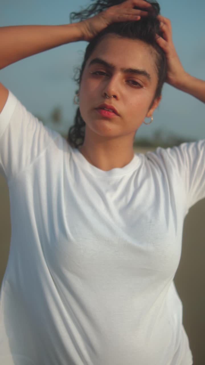 Indian woman in white t-shirt poses with hands in hair during golden hour on beach