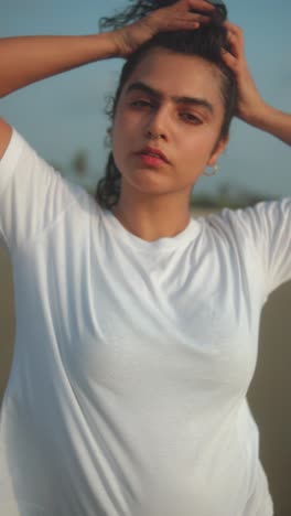 indian woman in white t-shirt poses with hands in hair during golden hour on beach