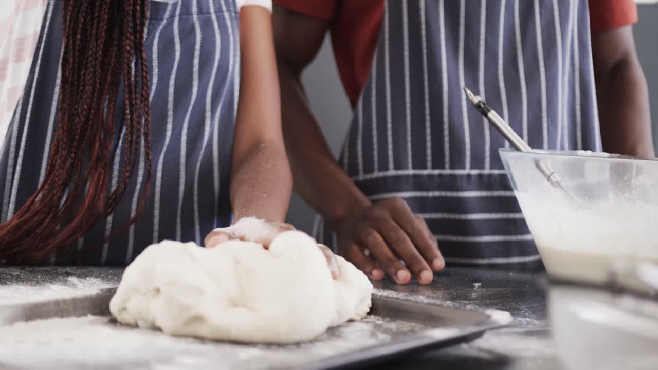Midsection of african american couple in aprons baking bread, kneading dough in kitchen, slow motion