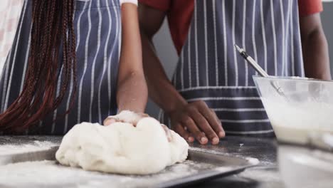 midsection of african american couple in aprons baking bread, kneading dough in kitchen, slow motion