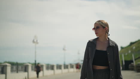 frau, die zuversichtlich auf einer brücke geht, sieht nach rechts, gekleidet in einem grauen hemd über einem schwarzen crop-top und sonnenbrille, im hintergrund eine verschwommene sicht auf eine andere person, die in der ferne steht