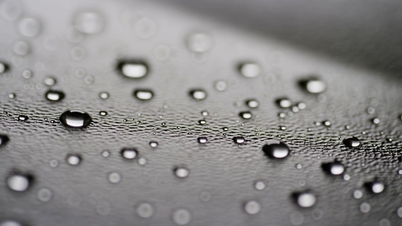 Close view of water droplets on dark textured surface with shallow depth, bright specular highlights, soft reflections, and directional light creating moody abstract detail